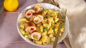 Visuel de roulés d'escalope de dinde milanaise Réghalal aux poivrons, posés sur des pâtes au citron et au parmesan dans un assiette blanche. Une fourchette en inox est posée dans l'assiette, le tout est sur un plan de travail de cuisine avec un torchon et un citron